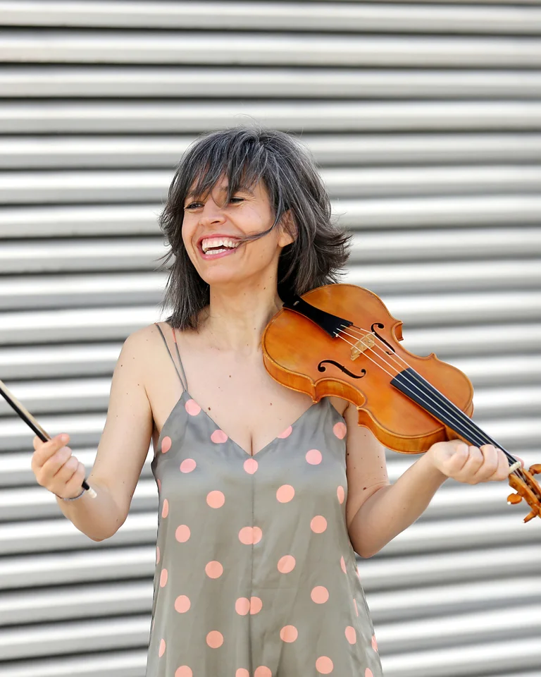 Frau mit schulterlangem Haar spielt eine Geige vor einem metallischen Hintergrund und lächelt. Sie trägt ein gepunktetes Kleid.