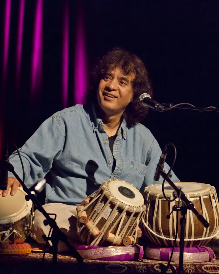 A musician playing tabla on stage, illuminated by purple lights. He's wearing a blue shirt and appears focused.