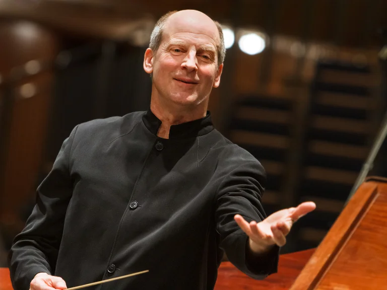 Werner Ehrhardt: Ein Dirigent in einem schwarzen Anzug dirigiert mit Ausdruckskraft ein Orchester in einem Konzertsaal.