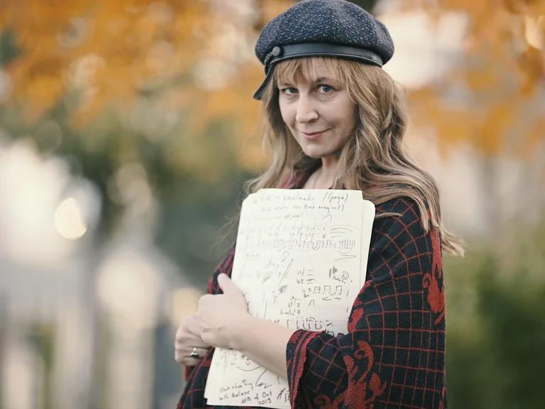 Eine Frau (Helena Winkelmann) im Freien hält Notenblätter in den Händen. Sie trägt eine Baskenmütze und einen karierten Mantel.
