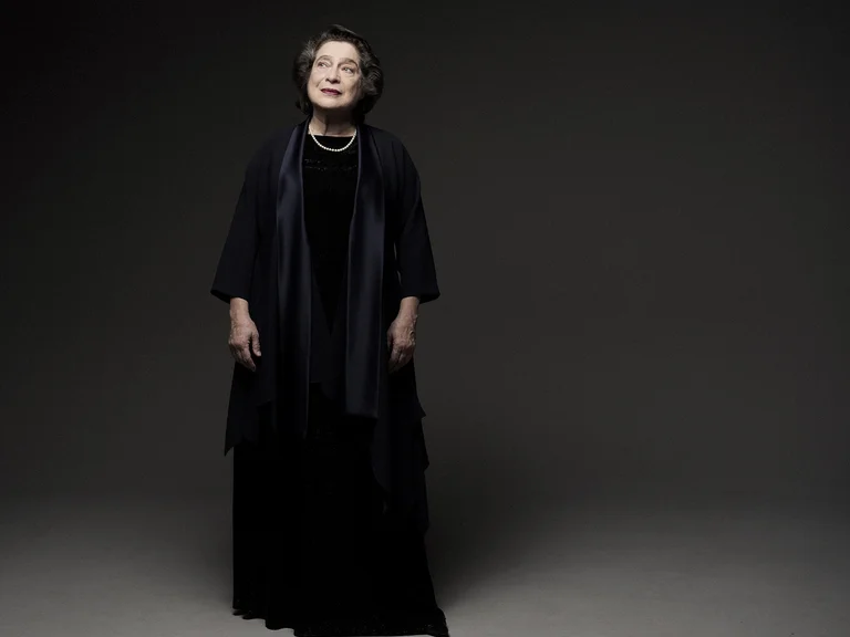 Eine Person in eleganter schwarzer Kleidung steht vor einem dunklen Hintergrund. Ihr Ausdruck ist gelassen, das Licht dezent.