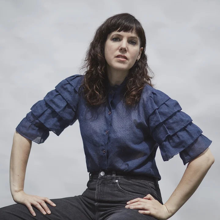 Anna Meredith is sitting in a blue blouse against a grey-and-white background. She is looking into the camera with a somewhat serious yet relaxed expression.