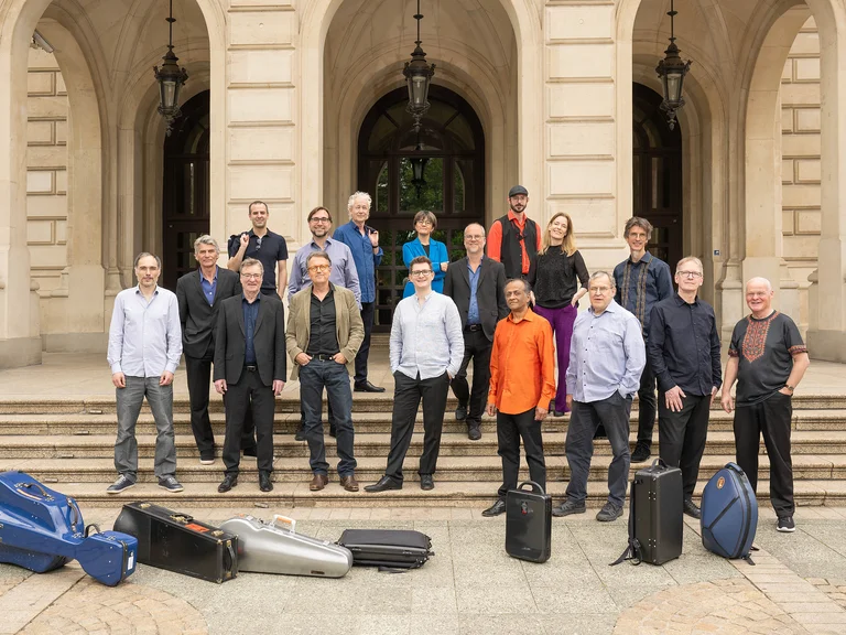 Eine Gruppe von Musikern (Ensemble Modern) steht vor einem historischen Gebäude mit Instrumentenkoffern auf den Stufen. Sie tragen verschiedene legere und formelle Kleidung.