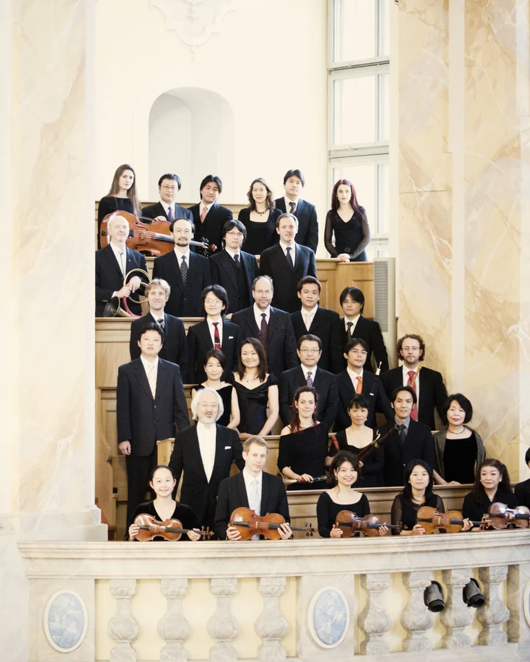 A choir in elegant attire stands on a marble staircase in a sunlit room within a historic building.