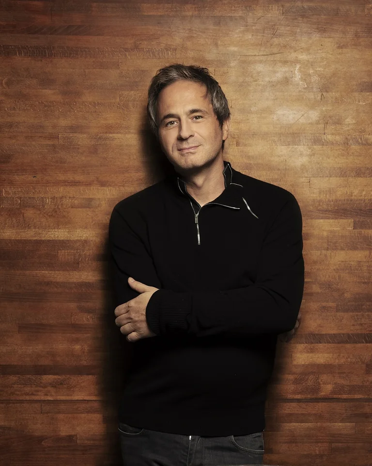 Eine Person in einem schwarzen Pullover steht vor einer Holzwand. Die Pose wirkt entspannt und selbstbewusst.