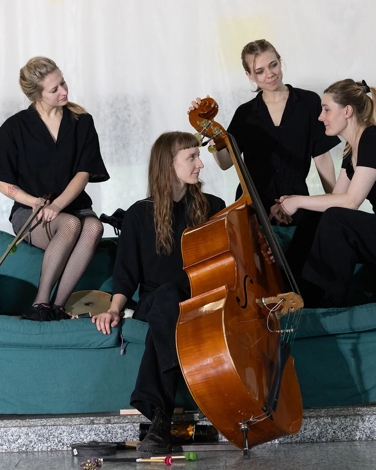 Vier Frauen in schwarzer Kleidung sitzen auf einem Sofa, eine mit Kontrabass, eine andere mit Querflöte. Musikalische Atmosphäre.