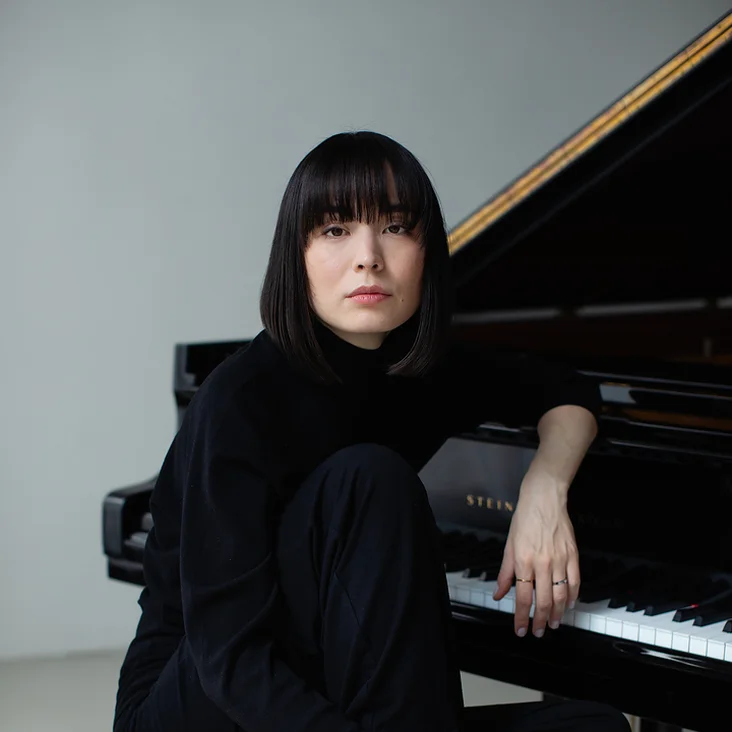 Eine Frau (Alice Sara Ott) in schwarzem Outfit sitzt barfuß auf Klavierbank vor einem Flügel, in nachdenklicher Pose mit ernstem Gesichtsausdruck.