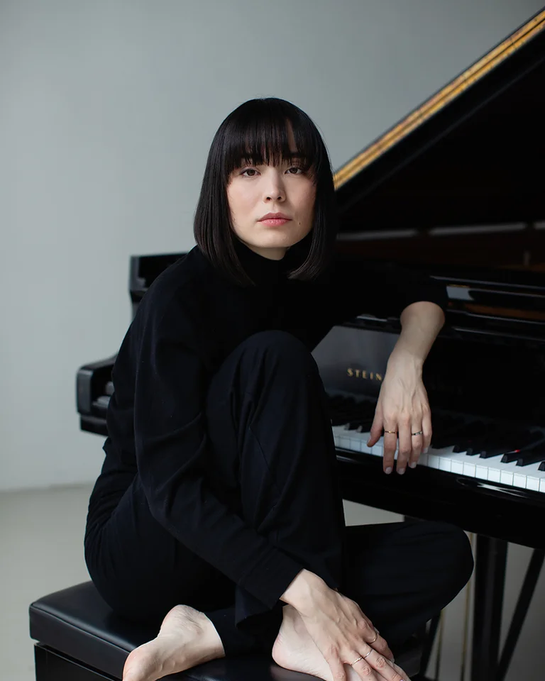 Eine Frau (Alice Sara Ott) in schwarzem Outfit sitzt barfuß auf Klavierbank vor einem Flügel, in nachdenklicher Pose mit ernstem Gesichtsausdruck.