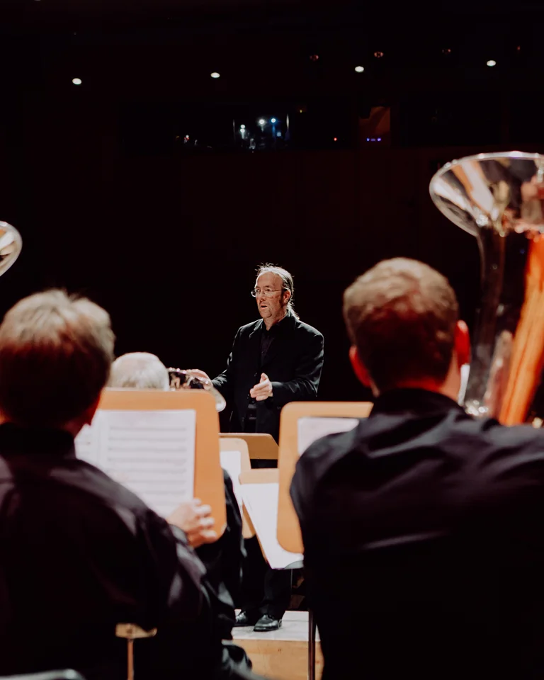 Eine Orchesterprobe mit Blechbläsern im Vordergrund, angeführt von einem Dirigenten. Die Musikerinnen und Musiker konzentrieren sich auf ihre Partituren.