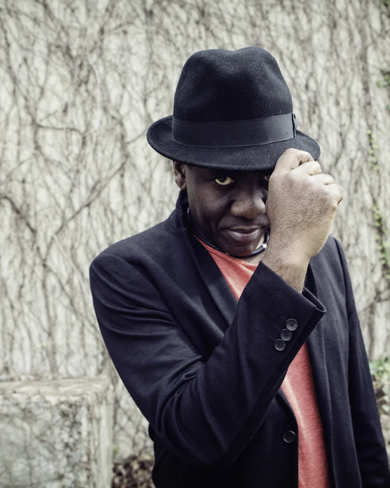 Ein Mann im schwarzen Mantel und Hut posiert draußen vor einer mit Efeu bewachsenen Wand.