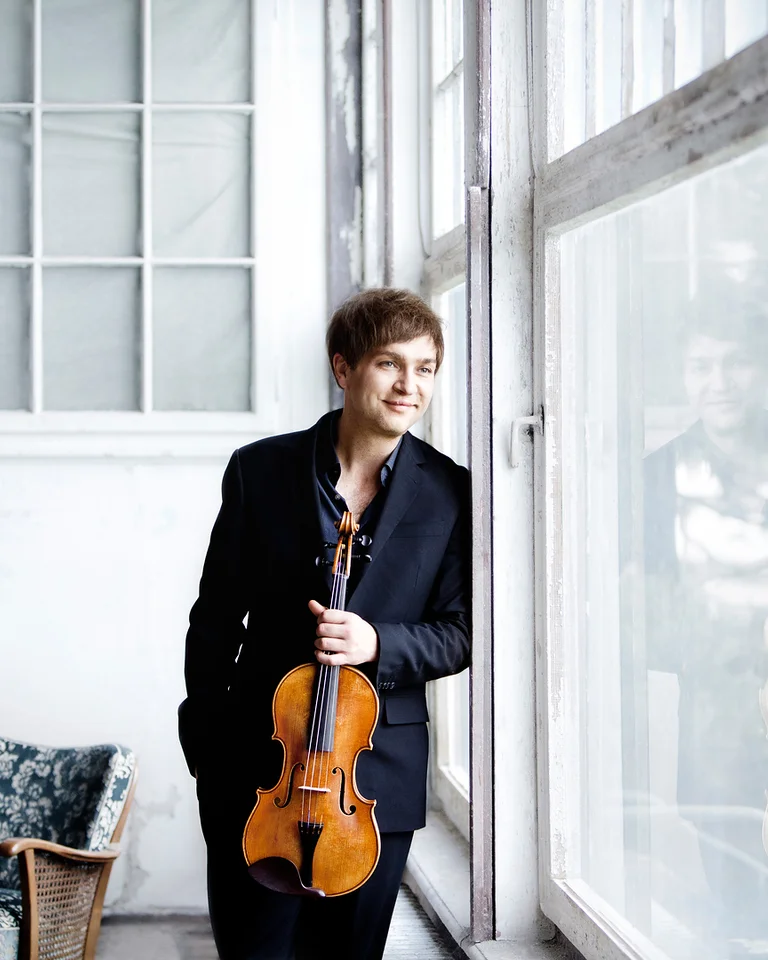 Eine Person (Nils Mönkemeyer) steht lächelnd an einem Fenster und hält eine Geige. Im Hintergrund ist ein Sofa zu sehen.