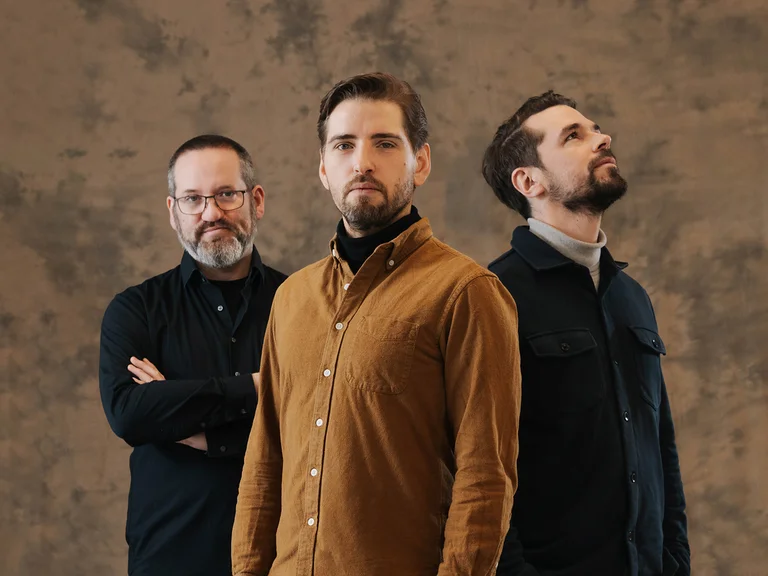 Drei Männer (Pablo Held Trio) in lässiger Kleidung stehen vor einem neutralen Hintergrund. Einer trägt ein braunes Hemd, die anderen schwarze Jacken.