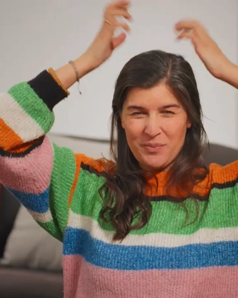 Eine Frau mit farbigem gestreiften Pullover schaut konzentriert und hebt die Hände, als ob sie gestikuliert oder tanzt.