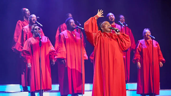 A group of singers in red choir robes (The Harlem Gospel Singers) performs passionately on stage, illuminated by blue lighting.