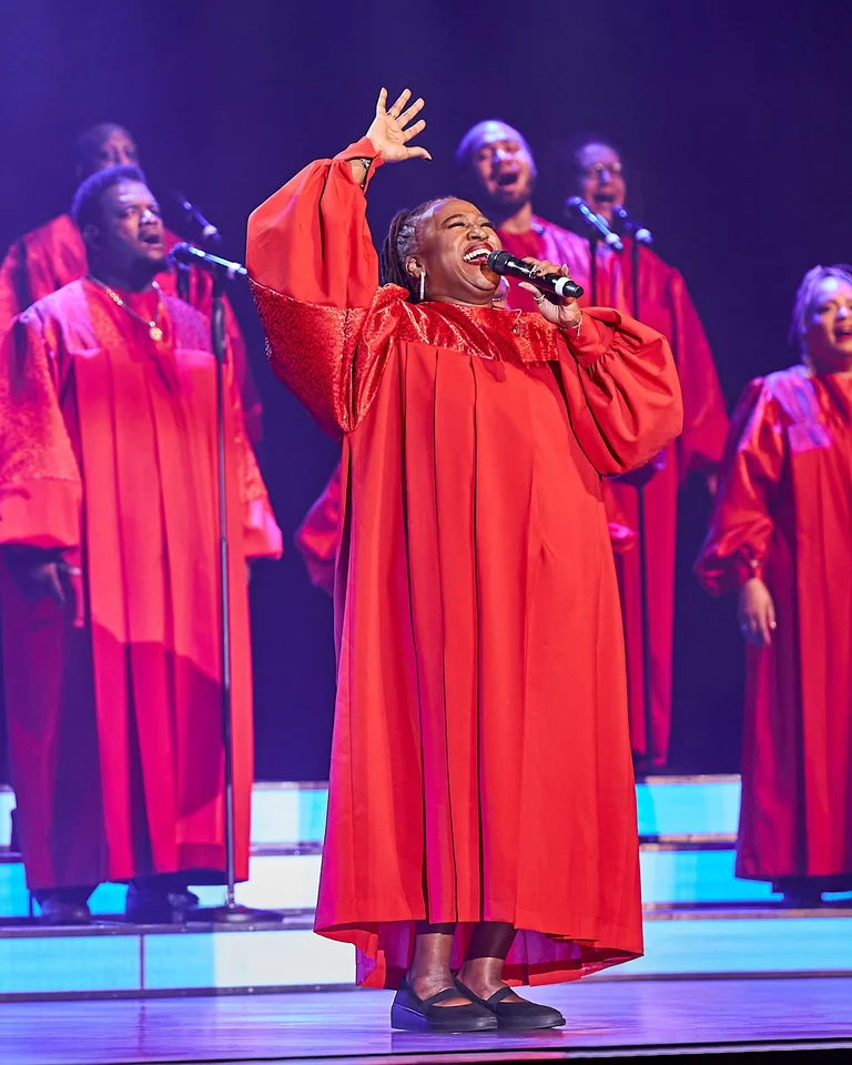 Eine Gruppe von Sängerinnen und Sängern in roten Chorkleidern (The Harlem Gospel Singers) singt leidenschaftlich auf einer Bühne, die in blauem Licht erhellt ist.