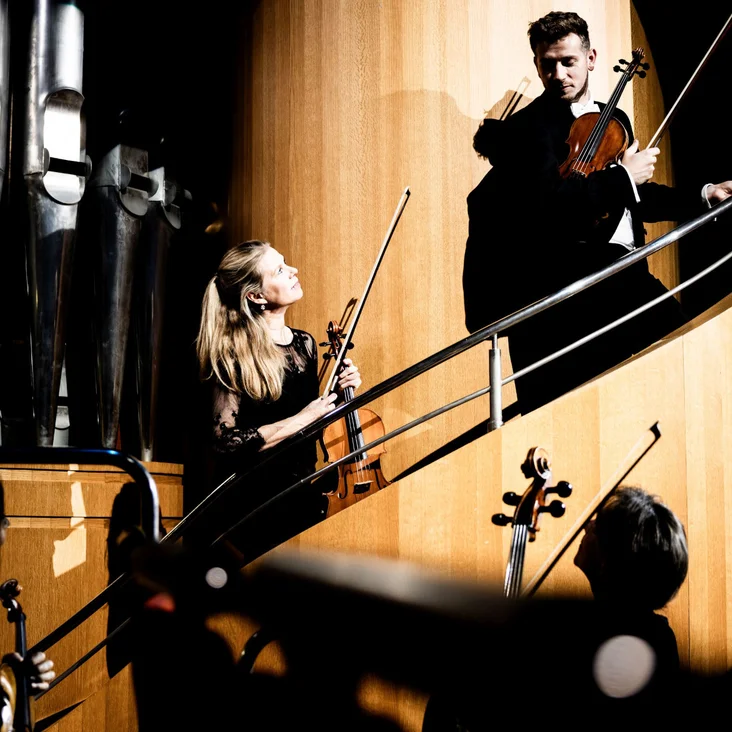 Ein Mann und eine Frau spielen Geige auf einer modernen Treppe in einem Konzertsaal. Lichter und Schatten sind intensiv.