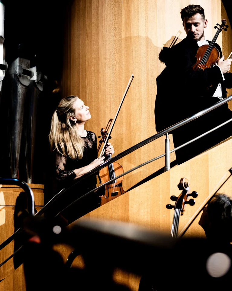 Ein Mann und eine Frau spielen Geige auf einer modernen Treppe in einem Konzertsaal. Lichter und Schatten sind intensiv.