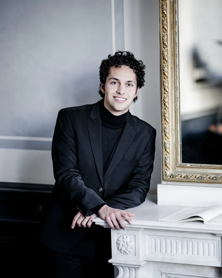 Ein Mann in einem schwarzen Anzug (Nikola Meeuwsen) steht lächelnd neben einem Klavier, das in einem Spiegel reflektiert wird.