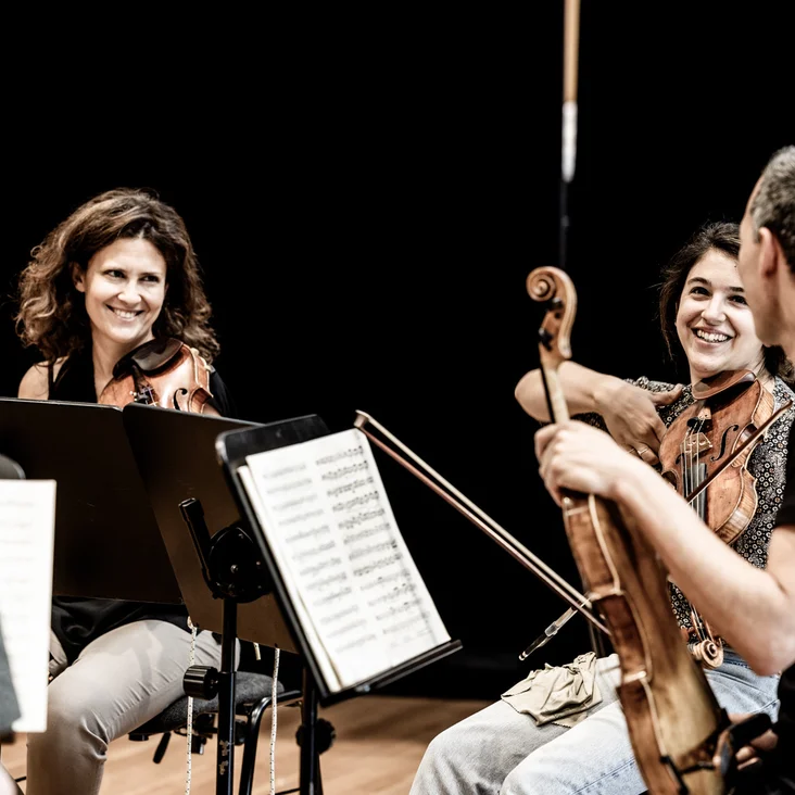 Eine Gruppe von Streichmusikern (Chamber Orchestra of Europe) sitzt auf der Bühne, lachend und entspannt, während sie sich auf ein Konzert vorbereiten.