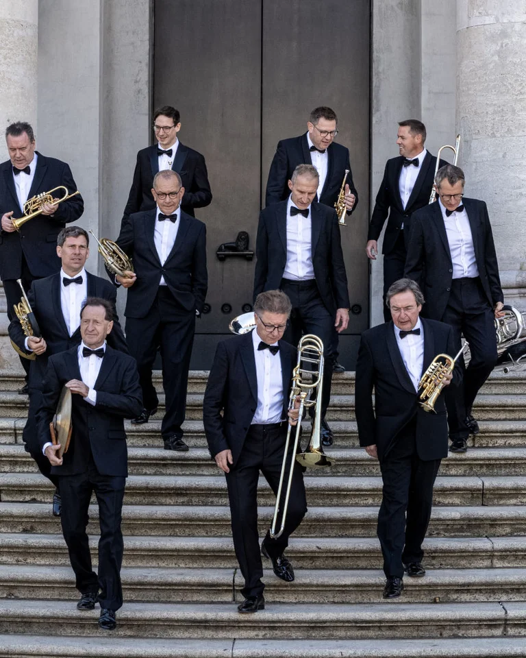 Eine zehnköpfige Blaskapelle in Smoking steigt Treppen vor einem großen Gebäude hinab, ihre Instrumente tragend.