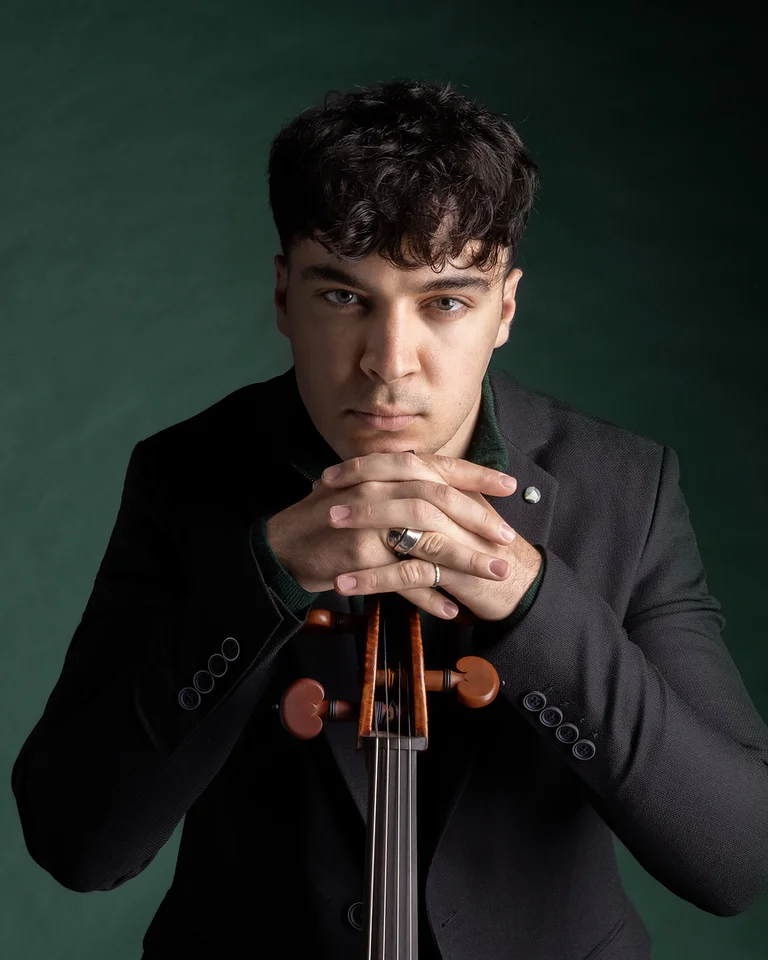 Ein Mann (Petar Pejčić) in einem schwarzen Anzug stützt seine Hände auf ein Cello. Er schaut ernst auf einem dunklen Hintergrund.