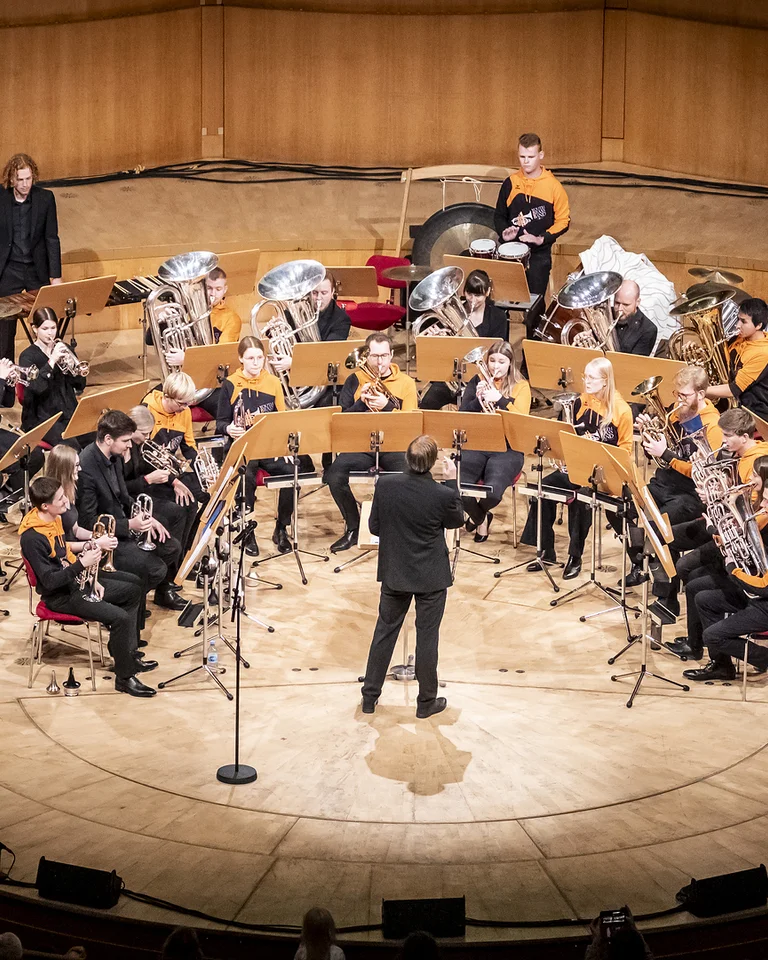 A large orchestra (Martin Schädlich mit Youth Brass Band NRW e.V.) performs in a concert hall. Many musicians with brass instruments, conducted by a person in the center.