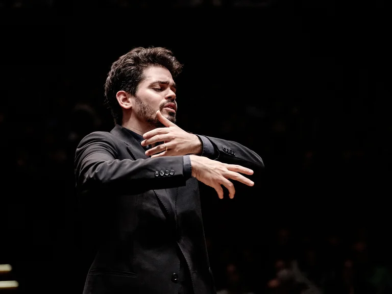 Ein Dirigent in formellem Anzug dirigiert leidenschaftlich ein Orchester, seine Hände und Gesichtsausdruck zeigen starke Emotionen.