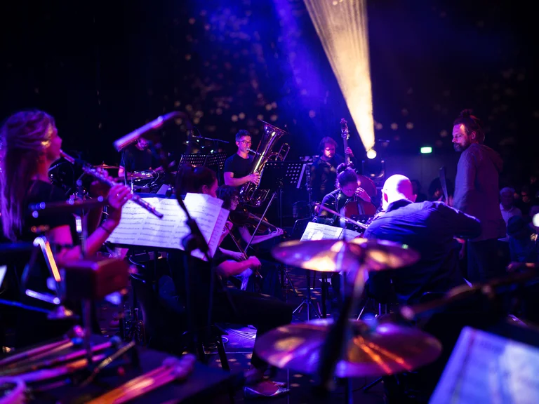 Ein Orchester (treppenhausorchester) spielt in einem stimmungsvoll beleuchteten Raum. Musiker mit verschiedenen Instrumenten erzeugen eine eindrucksvolle, harmonische Klangwelt.