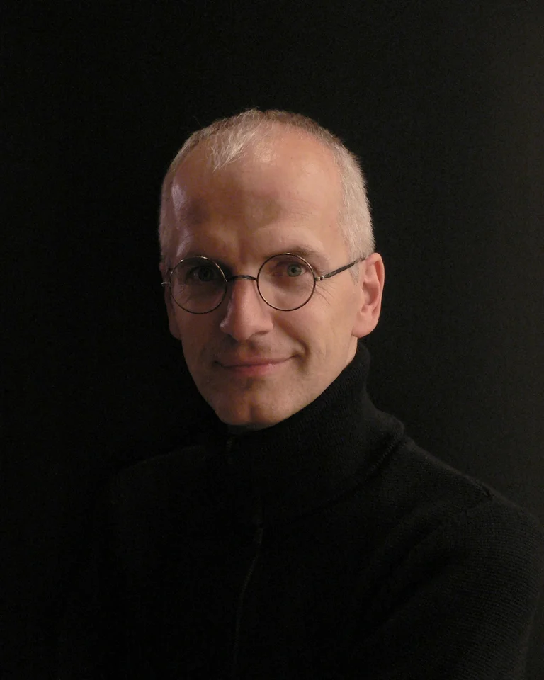 Portrait of a person wearing glasses and a black turtleneck against a dark background. The person is smiling softly.