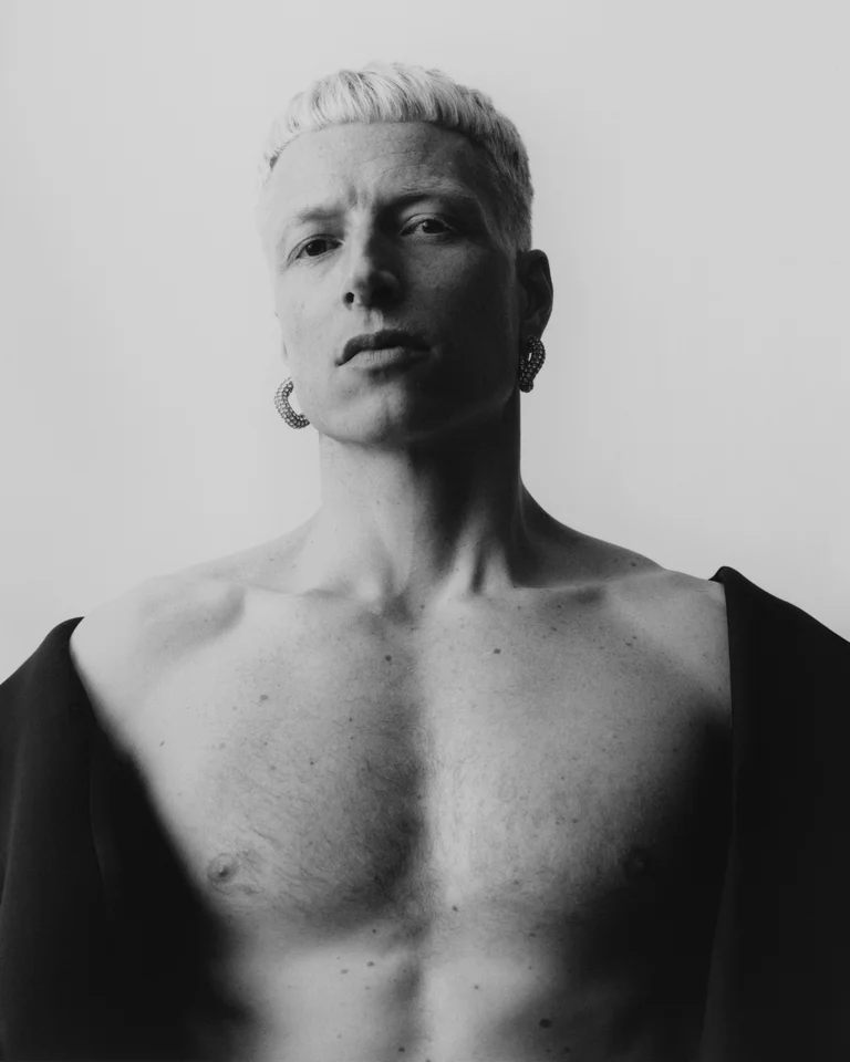 Schwarz-weiß Foto eines Mannes mit blondem Haar und offener Jacke. Er hat ein selbstbewusstes und nachdenkliches Auftreten.