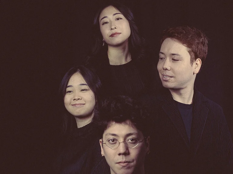 Vier Personen (Leonkoro Quartett) posieren vor einem dunklen Hintergrund. Sie tragen schwarze Kleidung und zeigen verschiedene Gesichtsausdrücke vor einer Kamera.