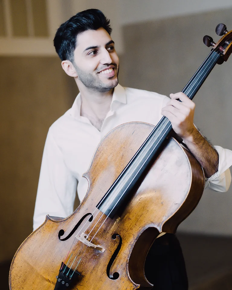 Ein lächelnder Mann (Kian Soltani) hält ein Cello in einem hellen Raum. Er trägt ein weißes Hemd und wirkt entspannt.
