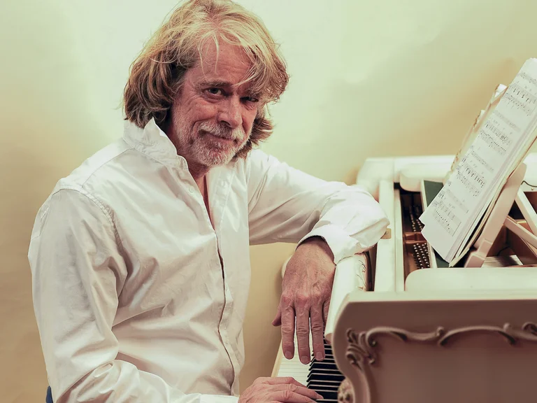 Mann mit langen Haaren und Bart sitzt am Klavier. Er trägt ein weißes Hemd und blickt freundlich in die Kamera.