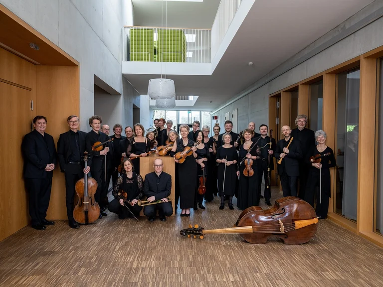 Eine Gruppe von Orchestermusikern (Freiburger Barockorchester) steht in einem modernen Flur. Sie halten verschiedene Streichinstrumente und sind in Schwarz gekleidet.