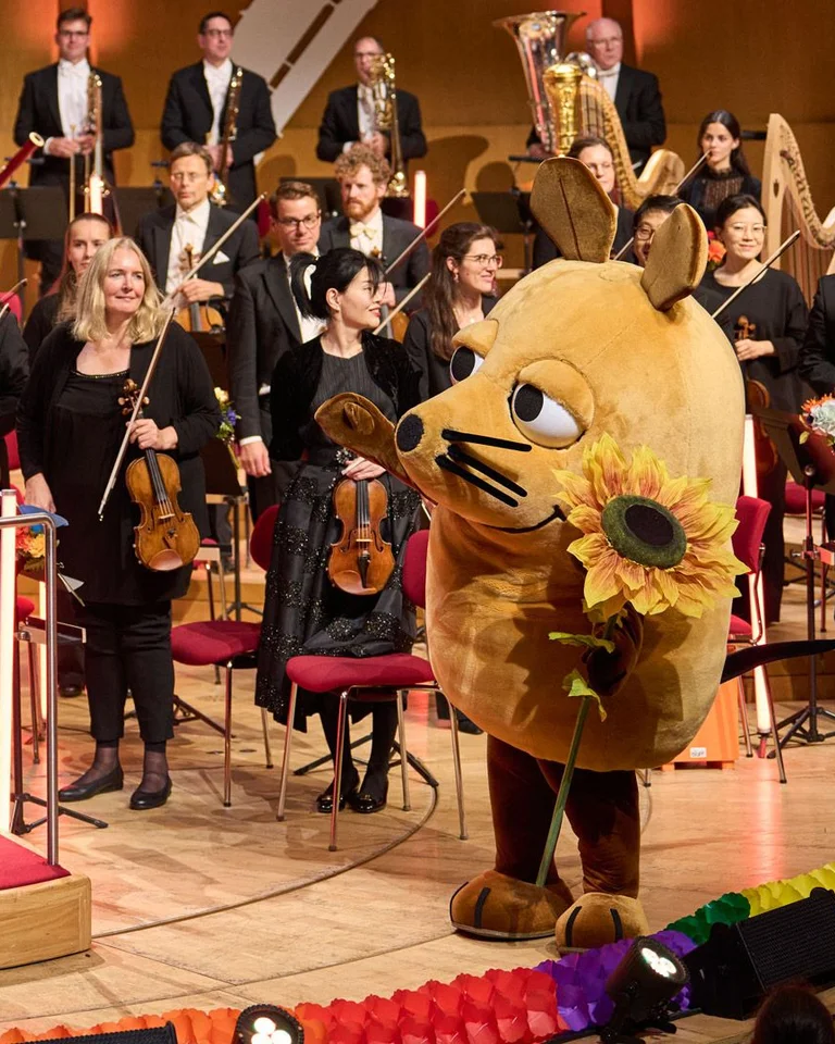 Ein Orchester mit einem Dirigenten auf der Bühne, daneben ein großes Maskottchen in Form einer Maus. Publikum applaudiert.