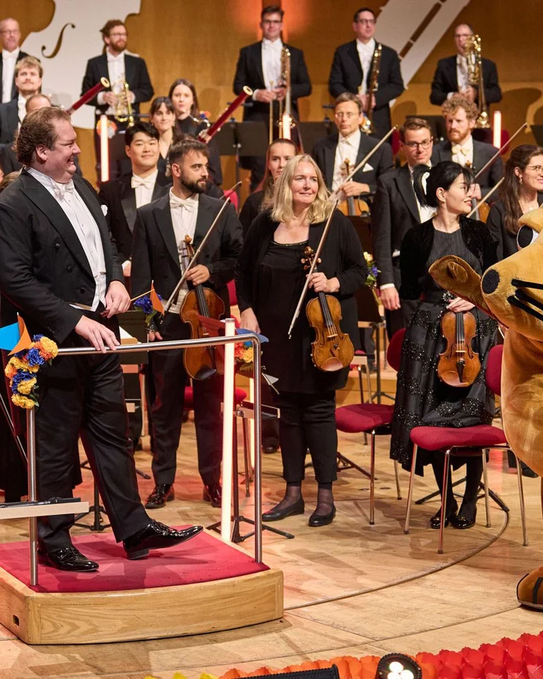 Ein Orchester mit einem Dirigenten auf der Bühne, daneben ein großes Maskottchen in Form einer Maus. Publikum applaudiert.