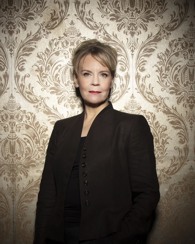 Eine Frau in schwarzer Kleidung steht vor einer kunstvoll gemusterten goldenen Tapete. Sie wirkt selbstbewusst und professionell.