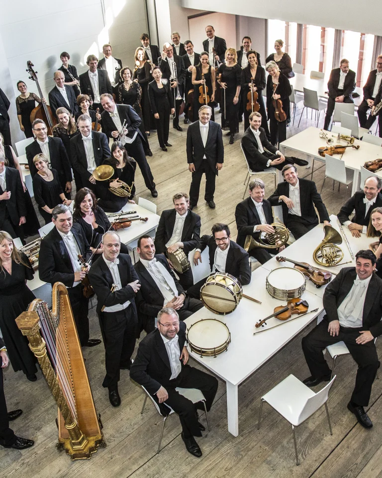 Ein großes Orchester in formeller Kleidung posiert in einem modernen Raum. Sichtbar sind verschiedene Instrumente wie Harfe und Schlagzeug.
