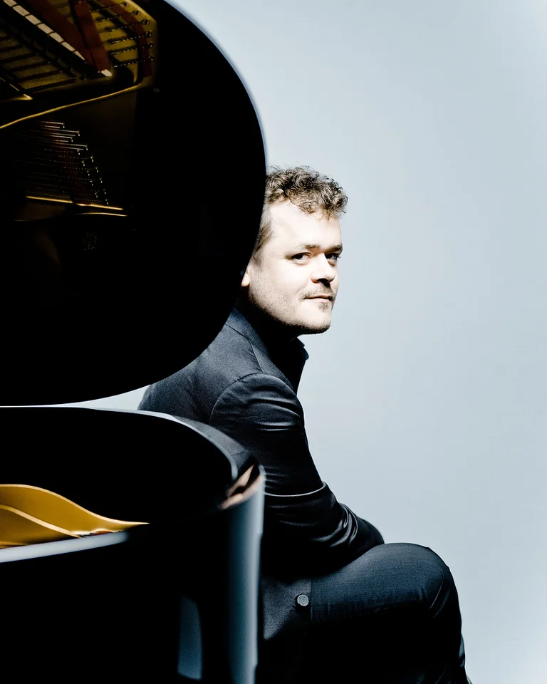 Ein Mann in einem schwarzen Anzug sitzt nachdenklich neben einem großen Flügel. Der Hintergrund ist schlicht gestaltet.