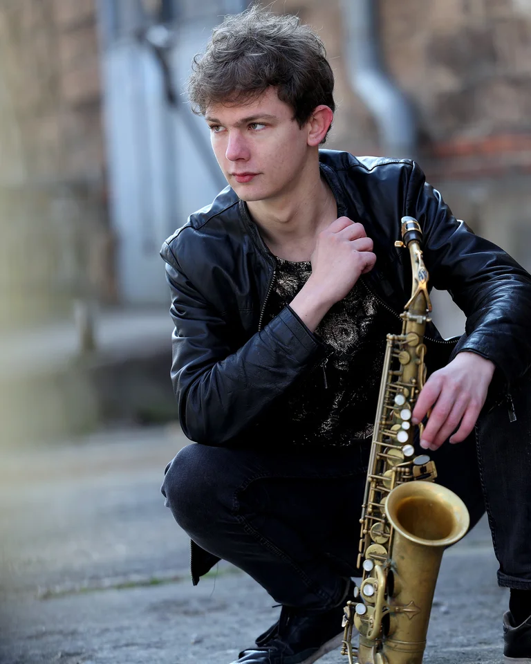 Jakob Manz in Lederjacke hockt mit einem Saxophon in einer urbanen Umgebung und blickt nachdenklich zur Seite.
