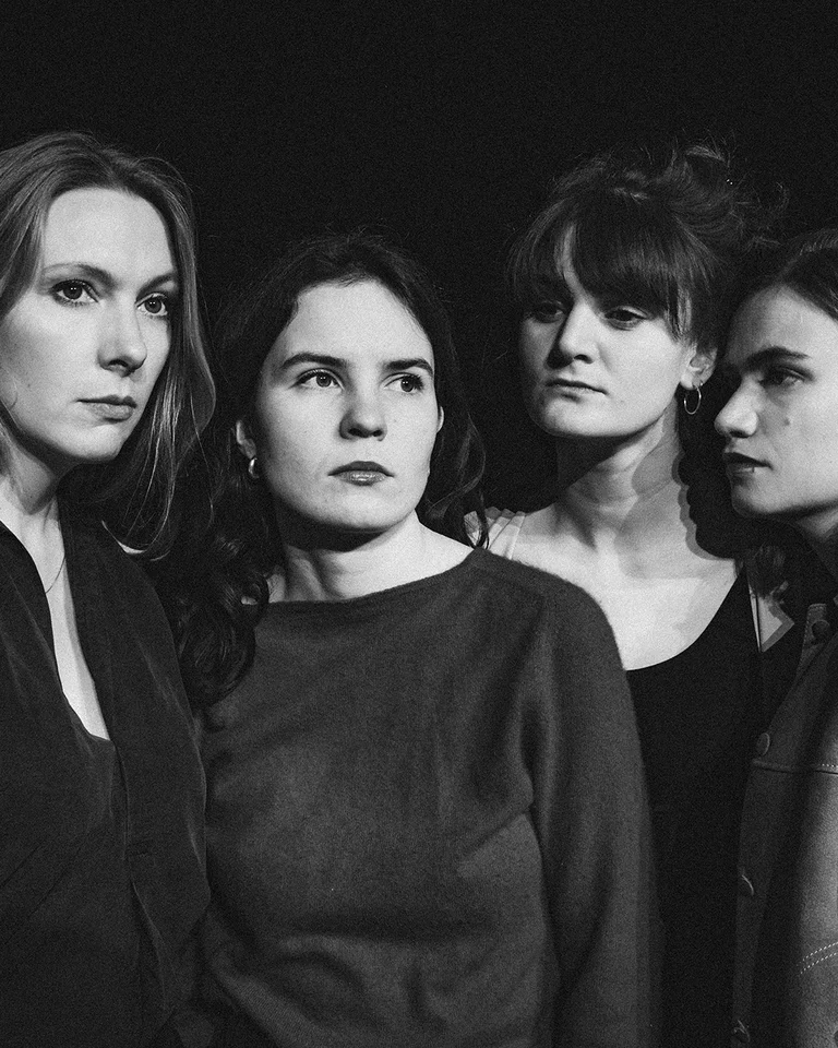 Schwarz-weiß Foto von vier Frauen, die stark und entschlossen wirken, in dramatischer Beleuchtung vor einem dunklen Hintergrund.