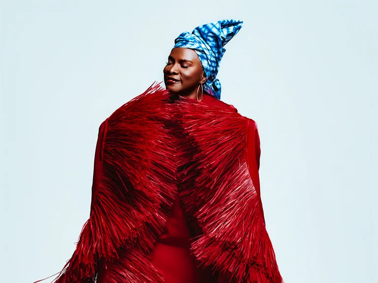 Frau (Angélique Kidjo) trägt ein voluminöses rotes Kleid mit langen Fransen und einen blauen, gemusterten Turban auf dem Kopf.
