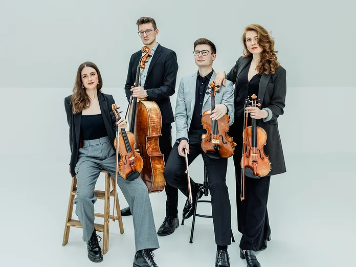 Four musicians in formal attire pose with their string instruments against a neutral background. Two are seated, two standing.