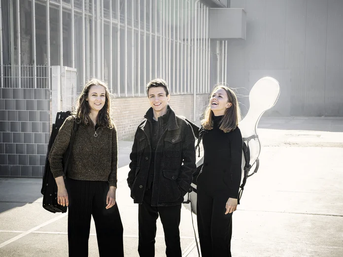 Drei Personen (Amelio Trio) stehen draußen in einer urbanen Umgebung, tragen schwarze Kleidung und lächeln. Sie haben Musikinstrumente dabei.