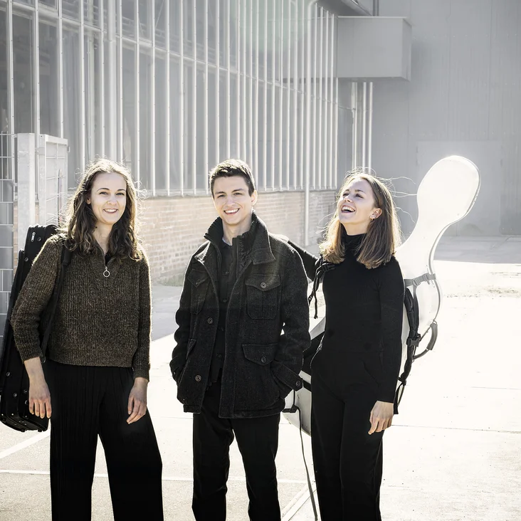 Drei Personen (Amelio Trio) stehen draußen in einer urbanen Umgebung, tragen schwarze Kleidung und lächeln. Sie haben Musikinstrumente dabei.