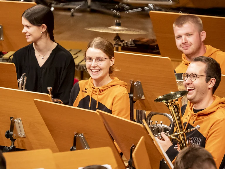 A group of musicians in orange hoodies (Youth Brass Band NRW e.V.) sit happily behind music stands, appearing to prepare for a performance.