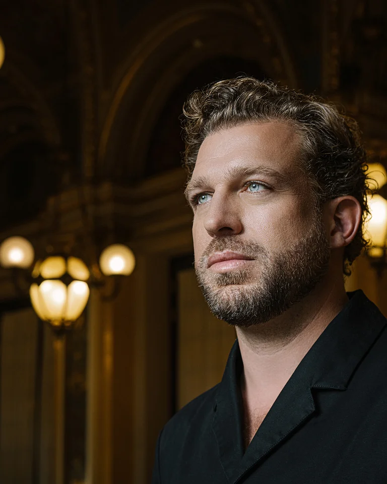 Ein Mann (Julian Prégardien) steht in einem eleganten Raum mit kunstvollen Leuchten im Hintergrund. Er trägt einen dunklen Anzug und blickt ernst.