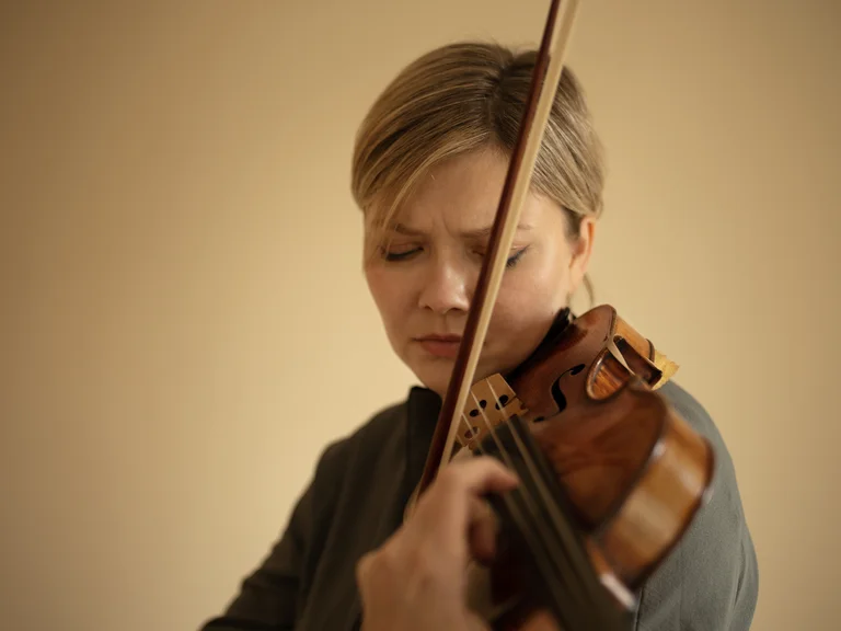 Eine Frau (Alina Ibragimova) mit geschlossenen Augen spielt konzentriert Geige, ihre Finger am Griffbrett und der Bogen in der Hand.