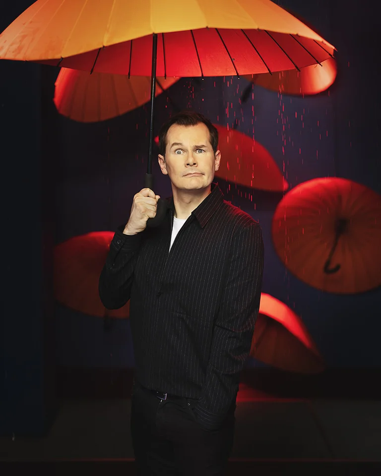 Ein Mann hält einen orangefarbenen Regenschirm vor einem dunklen Hintergrund mit weiteren orangefarbenen Schirmen.