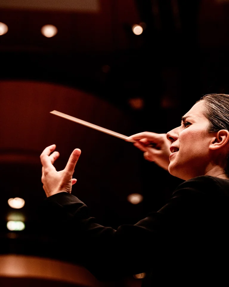 Dirigentin in einem Konzertsaal führt ein Orchester mit eleganten Bewegungen und einem Taktstock. Sanft beleuchtete Bühne.